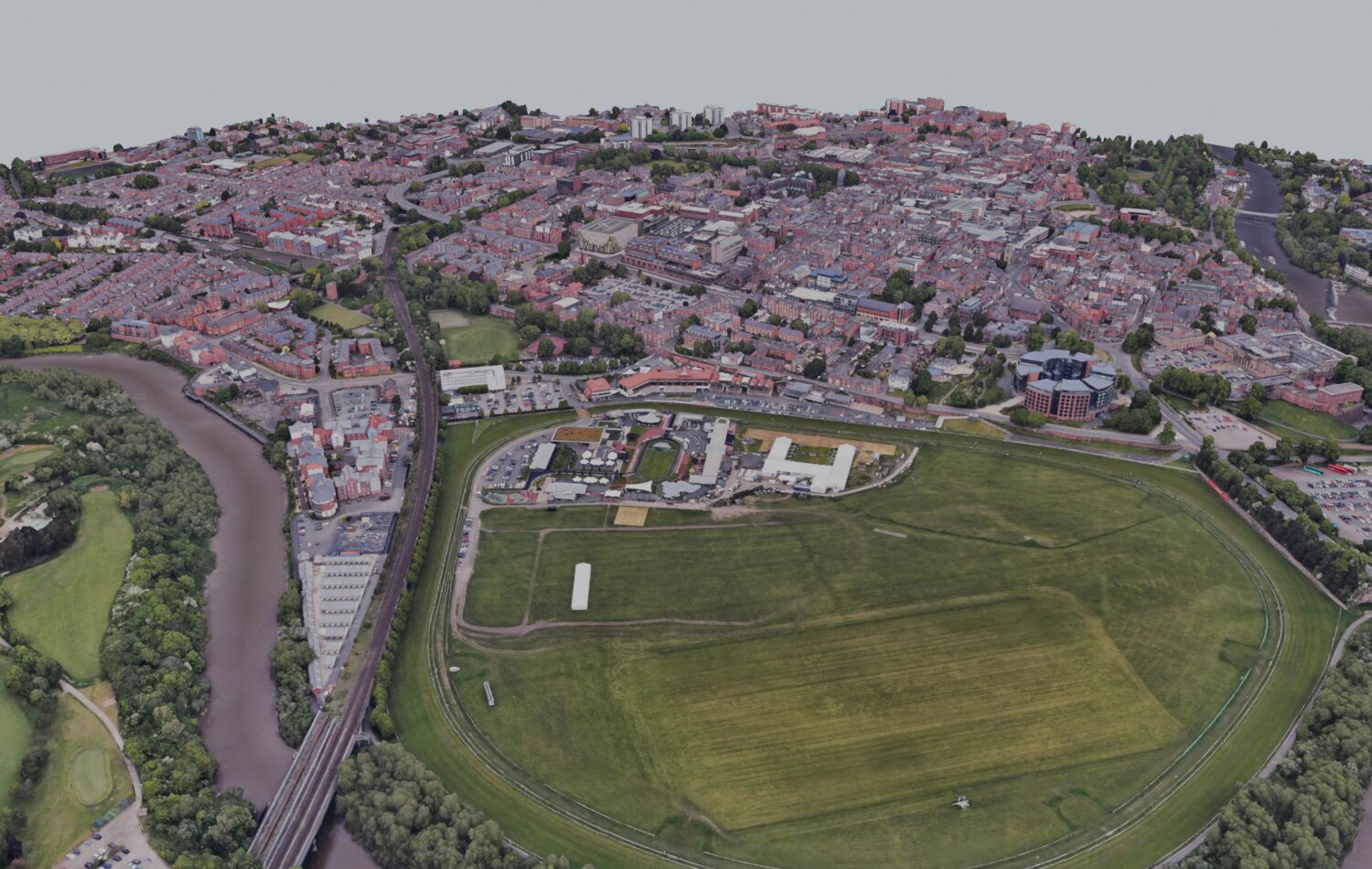 A wide oblique 3D view of Chester Racecourse, the Roodee, with the oval course and infield in the centre, the River Dee and railway viaduct to the left, and the city of Chester behind